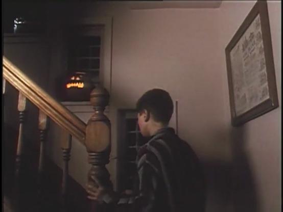 A boy is walking up a dimly lit staircase, while a glowing jack-o’-lantern can be seen in the window above.