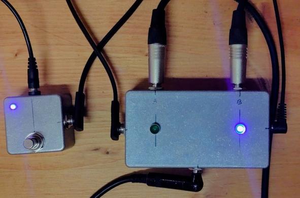 Photo of two pedals next to each other. One is small, has a footswitch and an LED. The other is larger, has two audio inputs with corresponding LEDs, and a single out. The two boxes are connected using a 6.3mm patch cable.