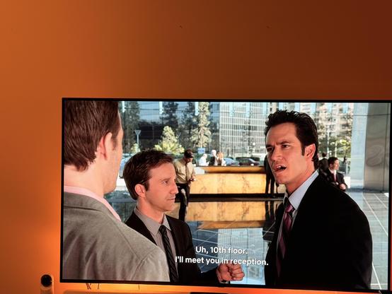 A scene from a television show or movie featuring three male characters in a modern office lobby. One character, dressed in a suit and tie, is speaking while gesturing towards another. The background shows a reception area and cityscape through large glass windows.