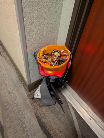 Photo of an orange bucket of candy, sitting on a 3 legged camping stool. There are assorted candies, mostly chocolates, including Reese's PB cups. Under the candy is a coiled LED light tube, and the white cord for it is snaking or of the bucket, under the door for power. The photo was taken from above, looking down, and the corners of the hallway walls of the hotel and a wooden hotel room door are around the bucket, which is sitting out of the way, in a corner on the outside of my door.