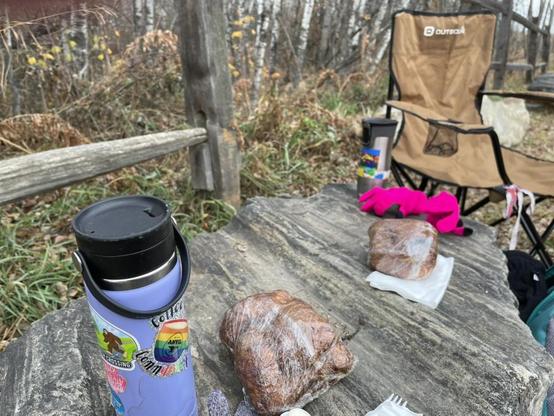 A camp chair, 2 travel mugs, and 2 cinnamon buns sit on top of a large rock.