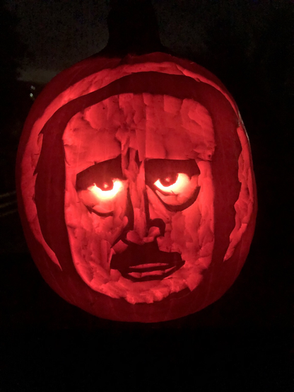 A pumpkin carving of a man with bushy eyebrows and bright glowing eyes that are cut fully from the pumpkin making them glow brighter. The rest of the carving is scored. The man has a groomed mustache, wild dark hair, and a pensive expression.