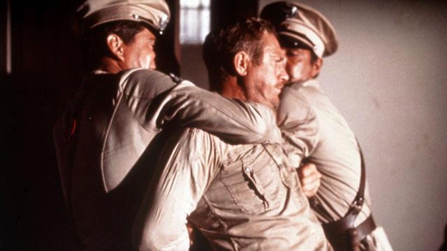 Drei Männer in hellen Uniformen mit Mützen halten einen Mann (Steve McQuen) in einem hellen Hemd fest. Die beiden Männer auf den Seiten greifen ihn an den Armen, er wird nach hinten gezogen und zurückgehalten. Die Szene spielt in einem Innenraum mit hellem Hintergrund.