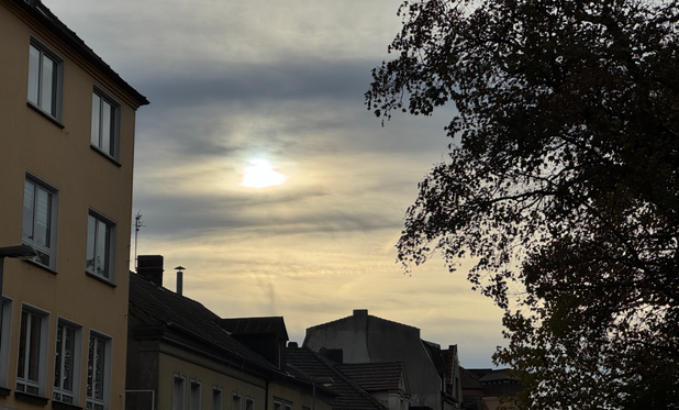 Das Bild zeigt eine städtische Szene bei bewölktem Himmel: Die Sonne scheint durch die Wolken und erzeugt einen markanten Lichteffekt. Im Vordergrund sind die Dächer und Fassaden von Wohnhäusern zu sehen, während im rechten Bildbereich ein Baumast mit Blättern ins Bild ragt.