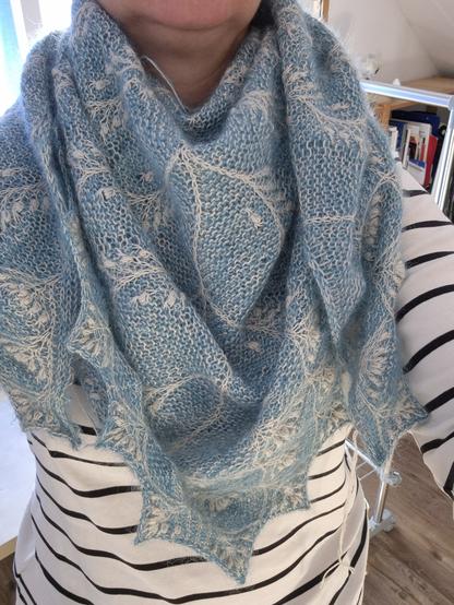 Eine Person trägt ein gestricktes Dreieckstuch um den Hals, darunter sieht man ein weißes Langarmshirt mit schwarzen Streifen. Kopf und Rest vom Körper sind nicht im Bild