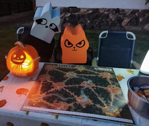 Orange cat cardboard box root meeple Halloween costume and grey racoon meeple cardboard box costume. With root board game on table with a fall pattern. Next to lit up pumpkin carved to show Enoch from over the garden wall. Ready to give it candy to trick or treaters.