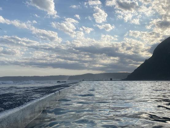 Looking southwards along the back wall at dusk while swimming in the main pool. The water catches the fading light and looks silky. The sky pale blue but full of white puffy clouds. Below it, the shadowed coastline stretches along the horizon.