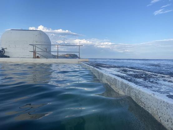 Looking northwards along the back wall of the baths towards the rounded white pump house building. The water is a deep greenish blue. Lower part of the sky along the ocean horizon has a bank of white cloud, the rest is blue.
