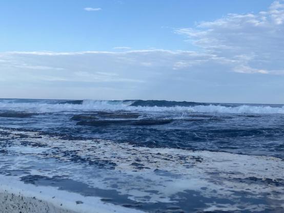 Looking over the back wall of the baths to the ocean horizon. Waves are breaking blue and white in the long shadow of the late afternoon. A thin skim of white clouds above then pale blue sky.