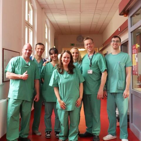 Eine Gruppe von sieben Personen steht in einem Klinikflur und schaut in die Kamera, unter ihnen Pfleger Ricardo und Ricarda Lang, MdB. Alle Personen tragen grüne OP-Kleidung. Der Korridor hat einen roten Boden, eine weiße Wand und eine Tür. An der Wand hängt ein gerahmter Fluchtplan. Die Person ganz links hebt den Daumen hoch.