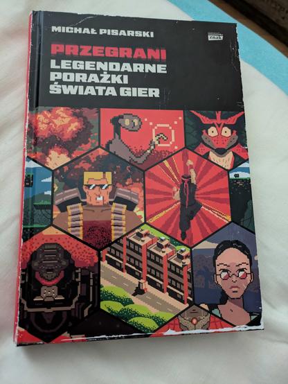 Zdjęcie książki "Przegrani. Legendarne porażki świata gier" autorstwa Michała Pisarskiego. Na okładce jest kilka postaci ze starych gier, a całość jest oprawiona w czerwono-biało-czarnej szacie graficznej, która wydaje się specjalnie uszkodzona.