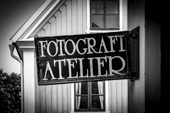 Vintage-style black and white photograph depicts a sign reading "FOTOGRAFI ATELIER" attached to a wooden building. The building has vertical siding and a window with curtains below the sign. Tree branches are partially visible on the left side.