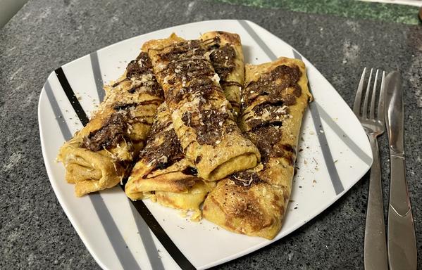a plate with four savoury crepes stacked on it. the crepes are rolled up in the shape of spring rolls or burritos, but are rather flat. they are topped with a dark sauce, and have been toasted in the oven. there is a knife and fork to the right of the plate.