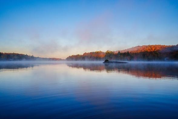A tranquil lake at dawn, surrounded by mist and colorful autumn foliage reflecting on the water.