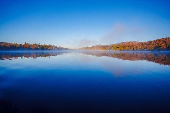 A panoramic view of a misty lake at dawn, with clear blue skies and colorful hills in the background.