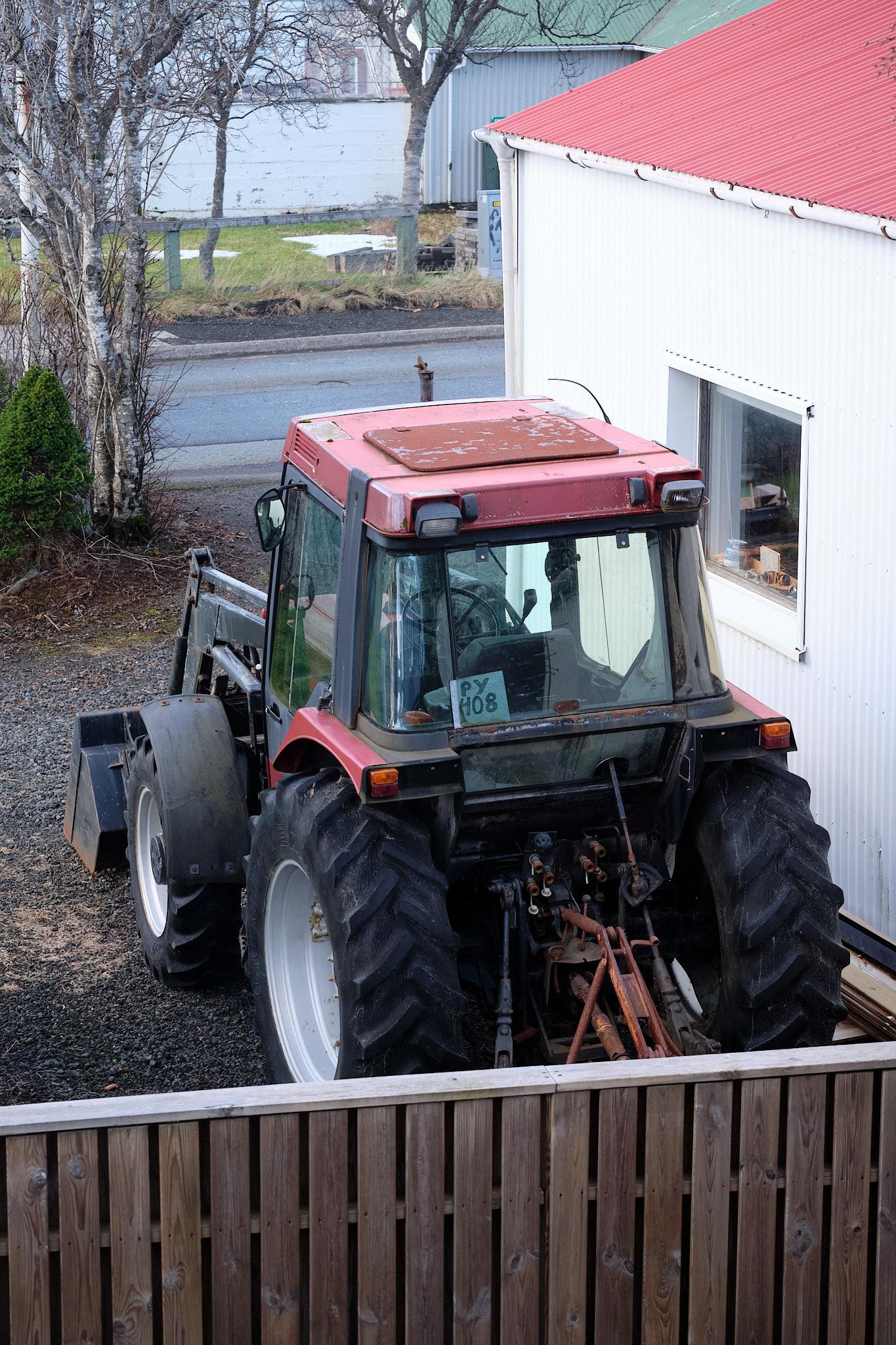 A photo of a very old and battered tractor parked on the other side of a fence next to a garage.