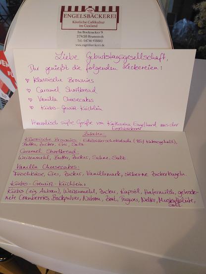 Auf einem großen, weißen Tortenkarton befinden sich zwei handgeschriebene Karten. Auf der oberen Karte steht folgender Text: Liebe Geburtstagsgesellschaft, Ihr genießt die folgenden Leckereien: klassische Brownies, Caramel Shortbread, Vanilla Cheesecakes, Kürbis-Gewürz-Küchlein. Himmlisch süße Grüße von Katharina Engelhard aus der Engelsbäckerei.

Auf der unteren Karte sind die Zutaten der einzelnen Kuchen aufgeführt.
Die klassischen Brownies bestehen aus Edelbitterschokolade mit 85% Kakaogehalt, Butter, Zucker, Eiern und etwas Salz.
Das Caramel Shortbread enthält Weizenmehl, Butter, Zucker, Sahne und Salz.
Die Vanilla Cheesecakes bestehen aus Frischkäse, Eiern, Zucker und Vanillemark.
Die Kürbis-Gewürz-Kuchen enthalten folgende Zutaten: Kürbis aus meinem Garten, Weizenmehl, Zucker, Rapsöl, Hafermilch, getrocknete Cranberries, Backpulver, Natron, Zimt, Ingwer, Nelke, Muskatblüte und Salz.

Wer Leckereien für einen bestimmten Anlass vorbestellt, bekommt selbstverständlich eine Übersicht der verwendeten Zutaten.