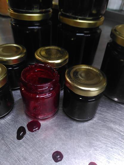 Auf einer Arbeitsfläche aus Metall stehen verschieden große Konfitüregläser mit goldenen Deckeln. Hier hinein habe ich Schlehen-Apfel-Konfitüre gefüllt. In der Mitte der kleinen Gläser steht eins, das nur noch Reste der Konfitüre enthält, da es mir leider umgefallen und ausgelaufen war. Es steht noch zwischen den anderen Gläsern, um zu zeigen, wie tief dunkelviolett die Konfitüre geworden ist.