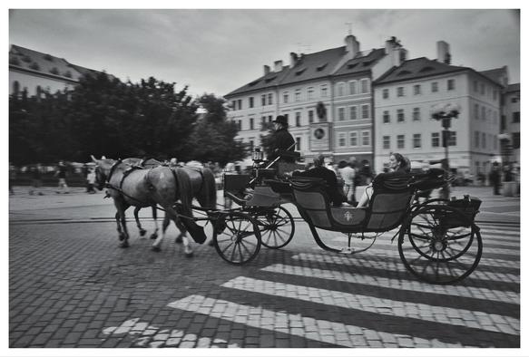 A black and white photograph showing a horse-drawn carriage moving from left to right across a cobblestone street and a crosswalk. The carriage is pulled by two horses. A driver wearing a hat is seated at the front, and two passengers are seated behind. Historic buildings of Prague line the background, with the scene taking place outdoors on a slightly overcast day. The street is paved with cobblestone. Pedestrian crossing is painted white over the cobblestone, and the carriage is caught crossing it.