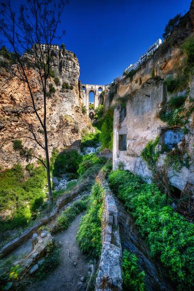 Hochformataufnahme einer engen, tiefen Schlucht, deren Felswände auf beiden Seiten hoch aufragen. Links eine nackte Baumkrone unter einem tiefblauen Himmel. Der Weg  und ein kanalisierter Bach sind im Vordergrund zu sehen. Rechts klammert sich ein verfallenes Steingebäude an die Felswand, teilweise überwuchert. In der Mitte, weiter hinten in der Schlucht, ist eine hohe Steinbrücke mit Bögen zu erkennen, die die Schlucht überspannt. Am oberen rechten Rand der Klippe sind helle Häuser einer Stadt sichtbar.