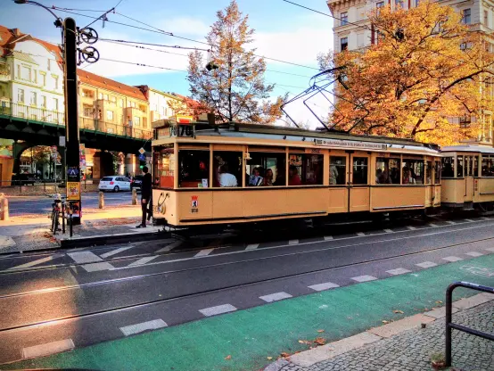 eine alte beige Straßenbahn fährt auf einer straße, wir sehen einen grünen fahrradweg, ein ubahn-viadukt, häuser, der himmel ist blau