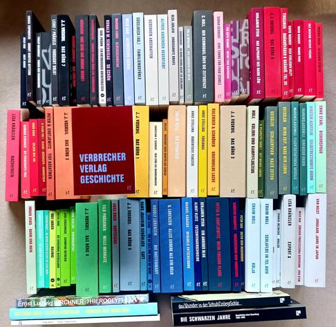 Foto des Buches, auf dessen dunkelrotem Cover sich neben dem kleinen Verlagslogo nur der Titel in drei Zeilen befindet – beides in einem Hellblau-Violett. Das Buch thront etwas erhaben über drei Reihen Büchern aus dem Verbrecher Verlag, die nach Farben sortiert sind. Wenn die Lektorin sich nicht verzählt hat, sind es insgesamt 79 Titel.