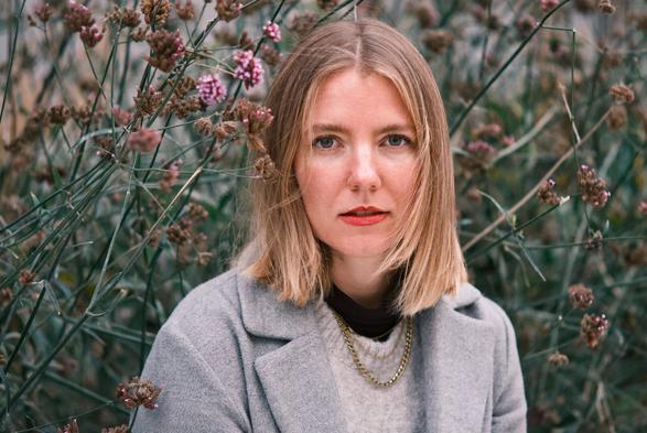 DE: Porträt einer blonden Frau mit grünen Augen. Sie trägt einen grauen Mantel, einen hellgrauen Pullunder und eine goldene Halskette. Ihr Blick geht direkt zur Kamera. Im Hintergrund sind grüne Stile von Pflanzen mit kleinen rosa zu erkennen. EN: Portrait of a blonde woman with green eyes. She is wearing a grey coat, a light grey slipover and a gold necklace. She is looking directly at the camera. In the background, green stems of plants with small pink flowers can be seen.