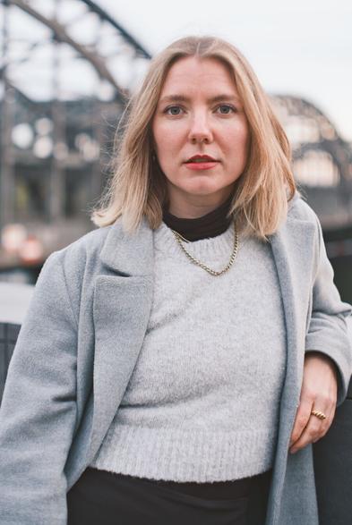 DE: Porträt einer blonden Frau mit grünen Augen. Sie trägt einen grauen Mantel, einen hellgrauen Pullunder und eine goldene Halskette. Ihr Blick geht direkt zur Kamera. Im Hintergrund sind Metallträger einer Brücke zu erkennen. EN: Portrait of a blonde woman with green eyes. She is wearing a grey coat, a light grey slipover and a gold necklace. She is looking directly at the camera. Metal girders of a bridge can be seen in the background.