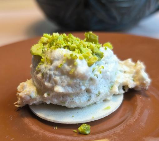 Eine weiße Kokosmakrone mit gehackten Pistazien darauf. Die Makrone ist ein Häufchen auf einer flachen Oblate, rechts und links quillt etwas über die Oblate hinaus. Die Makrone liegt auf einer braunen Untertasse, im Hintergrund steht eine dunkelblaue Keramiktasse.