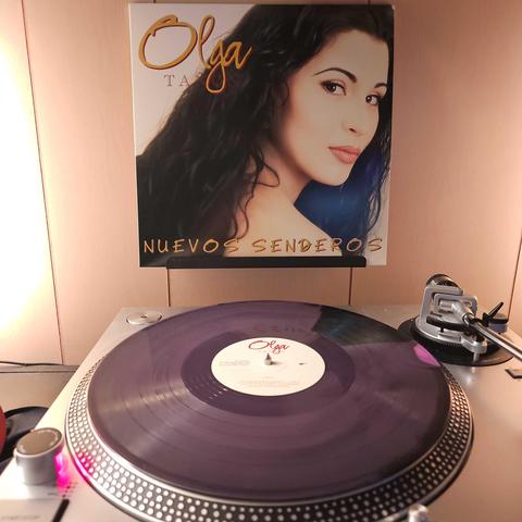 A a mostly purple with pink and black spots vinyl record sits on a turntable. Behind the turntable, a vinyl album outer sleeve is displayed. The front cover shows Olga Tanon from the shoulders up. She is looking back at the camera.
