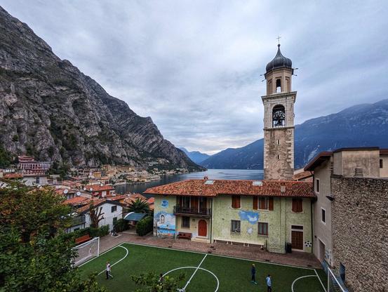 A picturesque village nestled between dramatic mountain cliffs and a serene lake. In the foreground, a small artificial turf football pitch is surrounded by a mix of traditional stone and pastel-coloured buildings with terracotta roofs. A few children are playing on the pitch. To the right, a tall stone bell tower with a dome and a cross rises above the village, adding a historic touch. The mid-ground reveals a cluster of tightly packed houses and buildings, leading down to the calm waters of the lake. The lake is bordered by steep, rugged mountains, partially shrouded in mist, creating a breathtaking and tranquil backdrop. The sky is overcast, enhancing the peaceful ambiance of the scene.