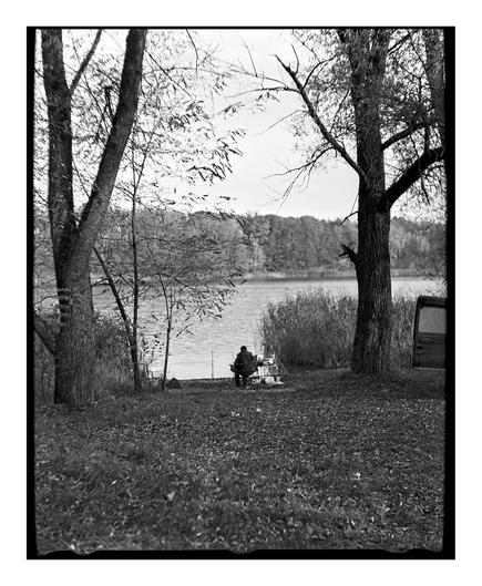 A black and white square film photograph of a person fishing. The person is sitting in a chair on the bank of a lake, framed between two large, leafless trees. The ground is covered in autumn leaves.