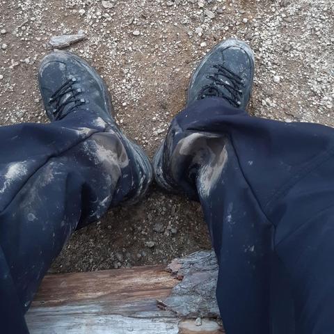 Photo of a person's leg and feet, the black hiking boots have muddy tips, and the black   pant legs show a lot of gray-brown mud.