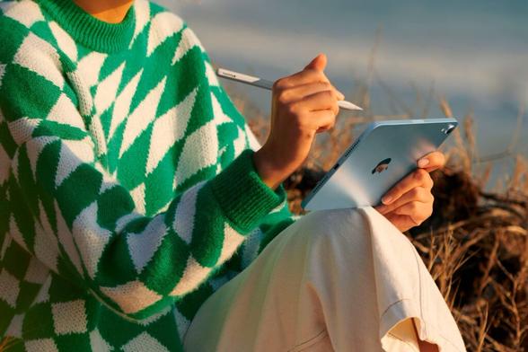 A person wearing a green and white patterned sweater is sitting outdoors, holding a blue Apple's 7th generation iPad mini in one hand and using a Apple Pencil Pro with the other. The iPad mini rests on their knee, with the background blurred to highlight the natural setting. The stylus is poised near the screen, suggesting active interaction with the device.