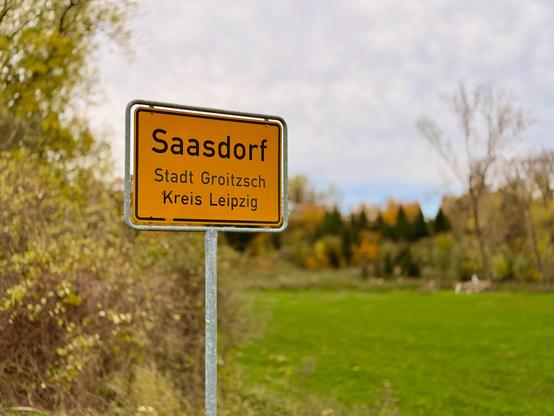 Deutsches gelbes Ortseingangsschild vor einem Hintergrund aus Büschen und Wiese. Darauf steht: Saasdorf, Stadt Groitzsch, Kreis Leipzig.
