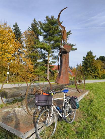 Effie sitzt im Fahrradkörbchen und das Rad lehnt an einer riesigen Metallstatue. Dabei handelt es sich um Charon, der die Toten über die Elbe bringt. Die Statue steht direkt auf dem Deich bei Mödlich nahe Lenzen. 
Im Hintergrund sind herbstlich gefärbte Bäume zu sehen.