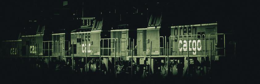 A row of Green Cargo locomotives parked in darkness. The image is split-toned with olive green shadow dominance and pale highlights. The branding is clear, especially the “green cargo” text which retains its green hue. The image conveys a heavy, systemic atmosphere with focus on structure, wheels, and railings.