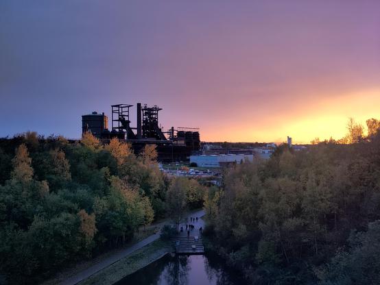 Ein Foto vom ehemaligen Hochofenwerk Phoenix-West bei einem Sonnenuntergang. Auf der rechten Seite des Bildes leuchtet ein flammend-oranger Sonnenuntergang und auf der linken ist der Himmel dämmernd blau-gräulich. Weiter vorne im Bild sind herbstliche Bäume, die einen kleinen Teich einschließen