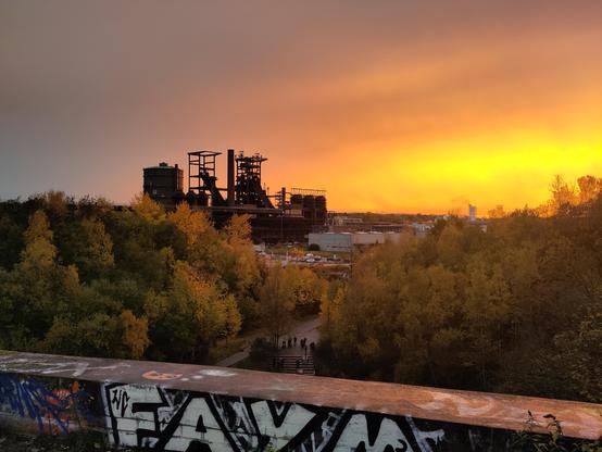 Ein Foto vom ehemaligen Hochofenwerk Phoenix-West bei einem Sonnenuntergang. Über den Himmel hinweg leuchtet ein flammend-oranger Sonnenuntergang. Etwas weiter vorne im Bild sind herbstliche Bäume. Unten im Bild, nahe der Kamera, ist eine alte graffitibedeckte Wand. Das auffälligste der Graffitis ist weiß auf schwarz und zeigt das Wort "FAYM"
