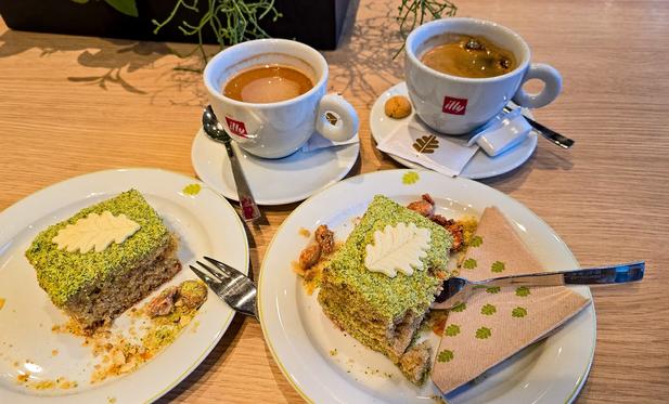 Zwei Tassen Kaffee und zwei Stücke Pistazienkuchen mit weißer Schokoladendekoration in Blattform stehen auf einem Holztisch. Auf den Tellern liegen Löffel, Gabeln und Servietten mit Blättermotiv.