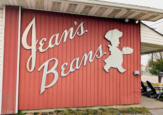 A building that has an orange-red vertical siding with sign letters in a vintage cursive style that reads "Jean's Beans" with a cut out of a jaunty silhouette of a chef in profile walking. Partially seen are Adirondack chairs and a pumpkin facing a street. (Eastern Blvd.)  01 Nov 2025