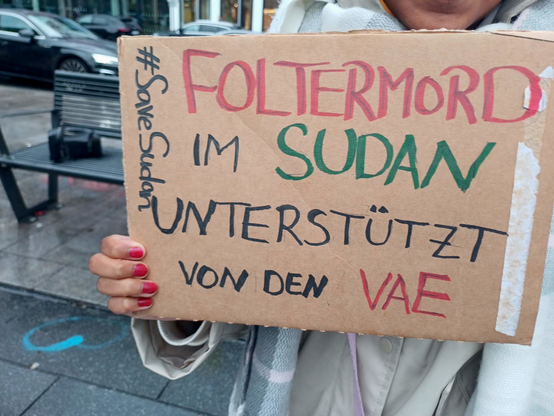 Eine Demonstrantin* hält ein Pappschild: "Foltermord im Sudan - unterstützt von den VAE - #SaveSudan"