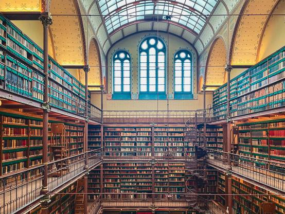 Wide-angle photograph of the Rijksmuseum Library in Amsterdam, showing multiple levels of bookshelves filled with colorful books, ornate railings, and tall arched windows letting in soft natural light. A spiral staircase connects the floors in this elegant, historic space.
