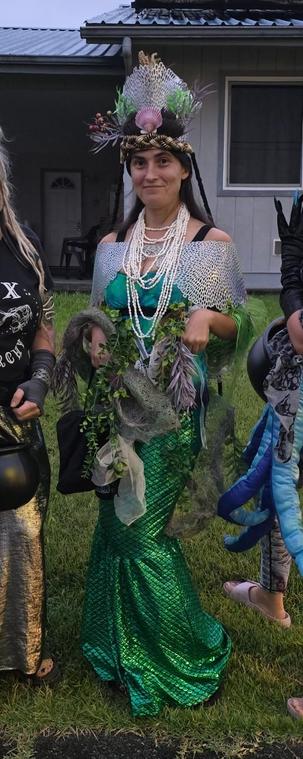 A gal in an overwrought sea queen costume with a mermaid skirt, crown, seashell necklaces, and water-plant boa