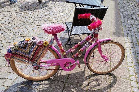 Ein Kinderfahrrad, das behäkelt wurde - der Sitz ist mit pinkfarbenem Fusselgarn bedeckt, der Rahmen in verschiedenen pink Tönen und gehäkelten Rosen. 

Der Gepäckträger / hintere Speichenschutz ist ein großes granny square, oben ebenfalls mit Rosen bedeckt. 

Die Reifen sind mit Strick bezogen, das Material sieht so echt aus, das man erst auf den 3. oder 4. Blick erkennt, dass es keine "echten" Reifen sind.

Das Rad steht auf der Straße, vor einem Stuhl, der vermutlich zu einem Straßencafé gehört.