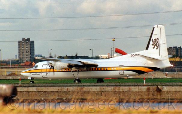 Side view of a high-winged, twin propellor-engined airliner moving from right to left along a runway, with flaps slightly extended from the rear of the wings, suggesting it is taking off..
The plane is white, with a yellow stripe outlined in black running along the body, turning down under the rear fuselage.
There is black text "Rotterdam" under the cockpit windows, the black registration on a white fillet at the front of the tail, and a rampant black lion on the white tail.
Long, yellowing grass lines the near side of a body of water that separates the camera from the runway, which is elevated several feet above the water.
Blocks of flats and trees line the horizon, with powerlines stretching across the fluffy grey clouds.
