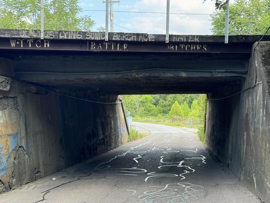 A photo of a railroad overpass with graffiti across the top that reads “WITCH BATTLE BITCHES”
