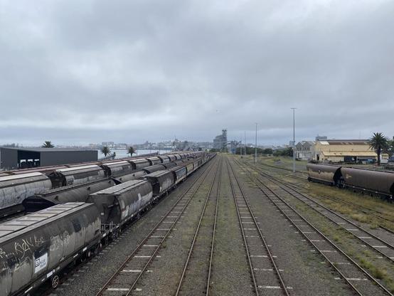 Looking down at and along the rail lines that run by the harbour near Carrington. Taken from a foot bridge that runs above the lines. Can see glimpses of harbour water on the left. And freight train carriages to the left and right of empty tracks.
