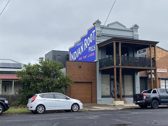 Light grey terrace style house with a big verandah with black wrought iron lace railings. On the side of the upper story, there's an ad for Dr Morse's Indian Root Pills in white on a blue background with "for the liver" written in red underneath.