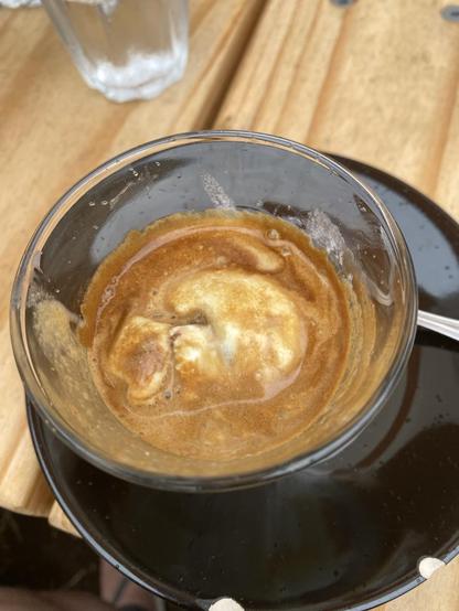 Glass with a shot of espresso poured over a scoop of vanilla gelato sitting in a black saucer on a light wooden table.